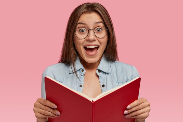 Photo d'une femme européenne intelligente qui tient un livre ouvert, se sent heureuse de lire une histoire romantique pour la fin, se sent excitée par un événement inattendu, porte une veste en jean et des lunettes rondes, se tient à l'intérieur