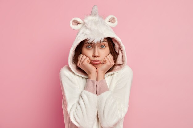 Photo d'une femme européenne mécontente garde les mains sous le menton, a l'air maussade, porte un costume de kigurumi, se sent seule, offensée par quelqu'un, isolée sur un mur rose. Fille malheureuse se tient à l'intérieur