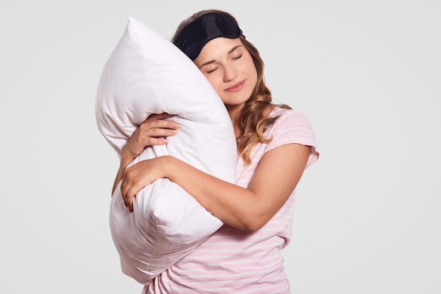 Photo d'une femme européenne avec une peau saine s'appuie sur un oreiller doux, porte un pyjama, des lunettes sur la tête, pose seule sur blanc, a l'air endormi. Gens, bonjour concept