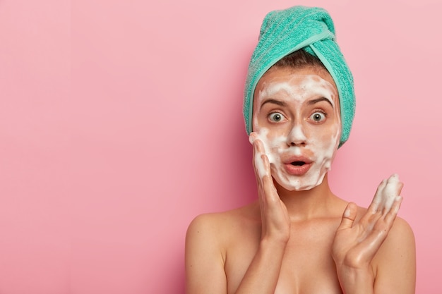 Photo d'une femme européenne surprise se lave le visage avec du gel mousse, veut avoir une peau bien soignée, se tient topless, porte une serviette enveloppée sur les cheveux mouillés, pose sur fond rose, espace libre de côté