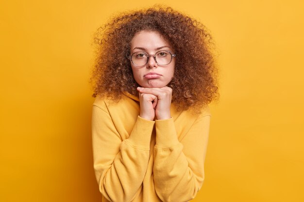 photo d'une femme européenne triste et déçue aux cheveux bouclés se sent contrariée garde les mains sous le menton les lèvres vêtues d'un sweat-shirt isolé sur un mur jaune se sent gênant et ennuyé