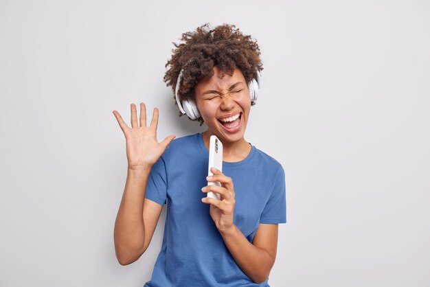 La photo d'une femme heureuse aux cheveux bouclés écoute sa musique préférée vêtue d'un t-shirt bleu décontracté utilise des écouteurs sans fil isolés sur fond blanc, pleine d'énergie, détient un microphone cellulaire comme