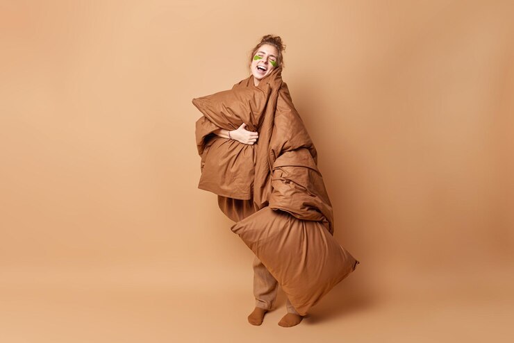Photo gratuit la photo d'une femme heureuse enveloppée dans une couverture douce tient un oreiller bénéficie d'une literie fraîche garde les yeux fermés rit joyeusement en allant dormir utilise des coussinets de beauté pose sur fond marron. bonne idée du matin