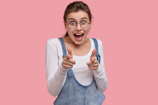 Photo de femme heureuse pointe avec les deux doigts à la caméra, a une expression joyeuse, sourit avec enthousiasme