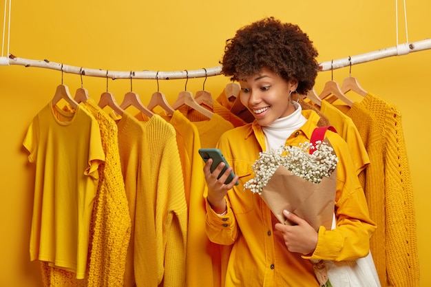 Photo d'une femme heureuse s'habille pour son premier rendez-vous, se tient près du porte-vêtements, reçoit des sms agréables sur un smartphone, tient un joli bouquet
