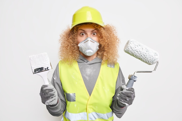 Photo d'une femme ingénieur bouclée et stupéfaite tenant un rouleau de peinture et un pinceau pour redécorer la maison vêtue de l'uniforme du bâtiment peint le mur intérieur du nouvel appartement