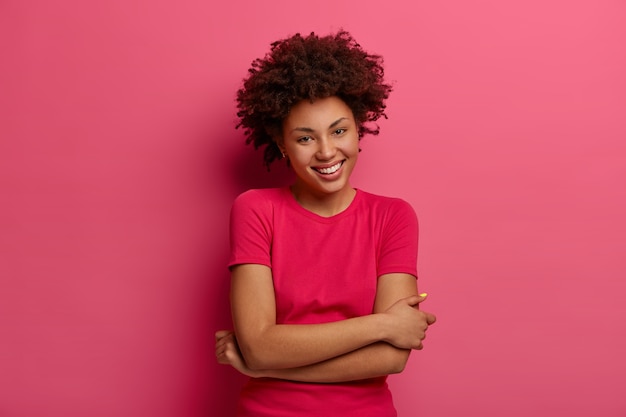 Photo d'une femme insouciante aux cheveux bouclés naturels, garde les mains croisées sur le corps, sourit agréablement, parle de manière décontractée, porte un t-shirt, profite d'une bonne journée, isolée sur un mur rose. Des sentiments heureux