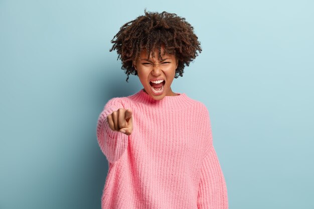 Photo d'une femme irritée aux cheveux bouclés, pointe directement la caméra, crie d'agacement