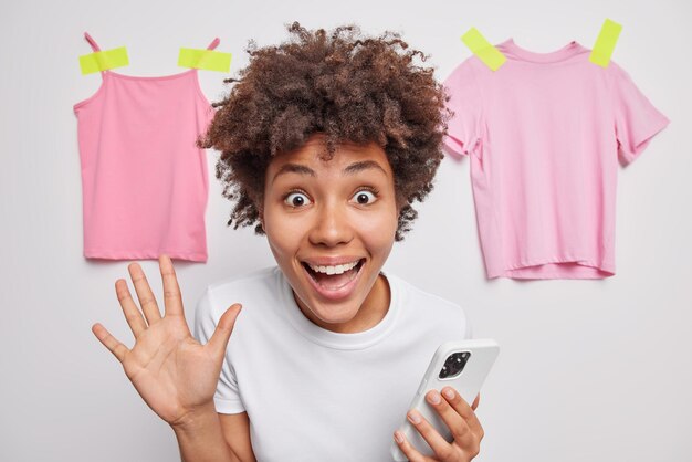 La photo d'une femme joyeuse aux cheveux bouclés se sent positive garde la paume levée utilise le téléphone pour discuter en ligne vêtue d'un t-shirt décontracté isolé sur un mur blanc avec des vêtements plâtrés réagit à des nouvelles incroyables