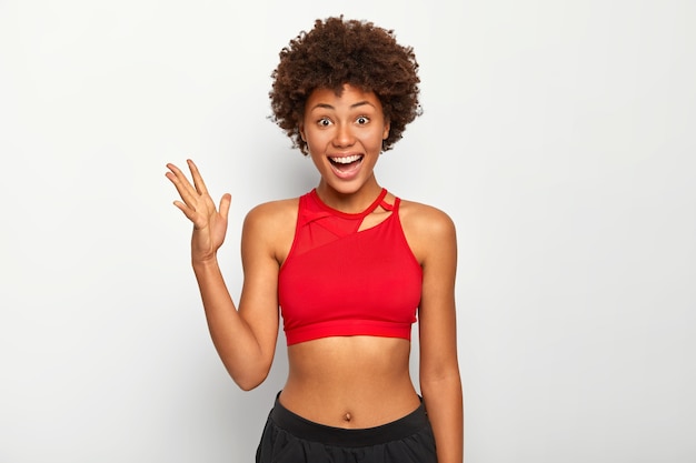 Photo d'une femme joyeuse à la peau sombre soulève la paume, porte un soutien-gorge de sport rouge, montre le ventre, a une coiffure afro, pose à l'intérieur sur fond blanc.