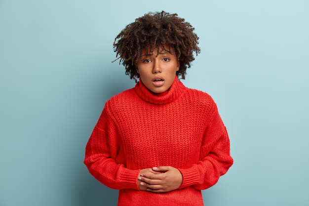 Photo gratuite photo d'une femme mécontente à la peau sombre touche le ventre, se sent mal après avoir mangé de la nourriture avariée, porte un pull rouge