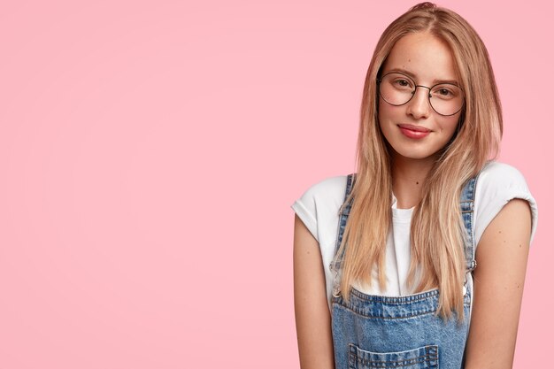 Photo d'une femme de race blanche ordinaire aux cheveux longs, a une expression positive timide, vêtue d'une tenue décontractée, se dresse sur un mur rose avec copyspace