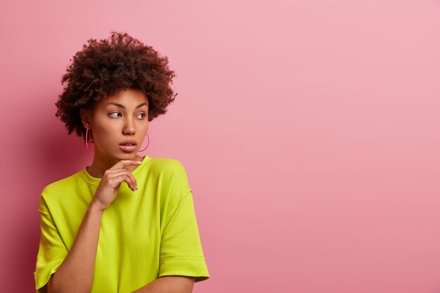 Photo d'une femme réfléchie avec des cheveux afro bouclés, garde la main sous le menton, a une expression pensive calme, porte un t-shirt vert décontracté, isolé sur un mur rose,