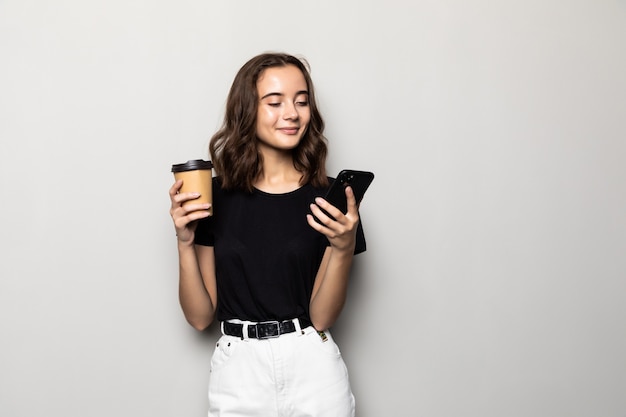 Photo de femme réussie en tenue de soirée debout avec smartphone et café à emporter dans les mains isolées sur mur gris