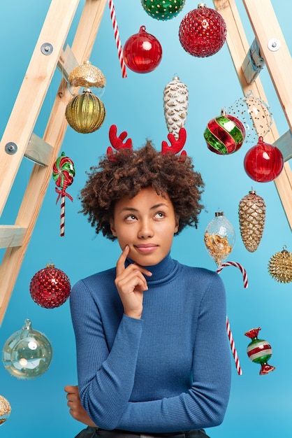 Photo d'une femme sérieuse à la peau sombre et aux cheveux bouclés pense quel cadeau acheter pour son mari le Nouvel An vêtu d'un col roulé décontracté et de cornes de renne entourées de boules brillantes accrochées à une échelle