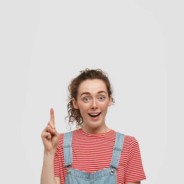 Photo d'une femme surprise excitée avec une peau tachetée de rousseur, pointe avec l'index, montre quelque chose d'étonnant vers le haut