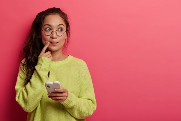 Photo d'une femme surprise pensive garde l'index près des lèvres, regarde de côté, pense au contenu du message, tient le téléphone mobile