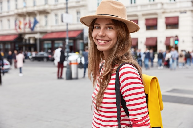 Photo gratuite photo de femme touriste joyeuse souriante se sent bien après une promenade à travers la ville, porte une coiffe élégante et un pull rayé