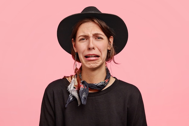 Photo d'une femme triste pleure comme le chagrin, serre les lèvres et a une expression faciale mécontente, porte un chapeau et un pull élégant, pose contre le mur rose