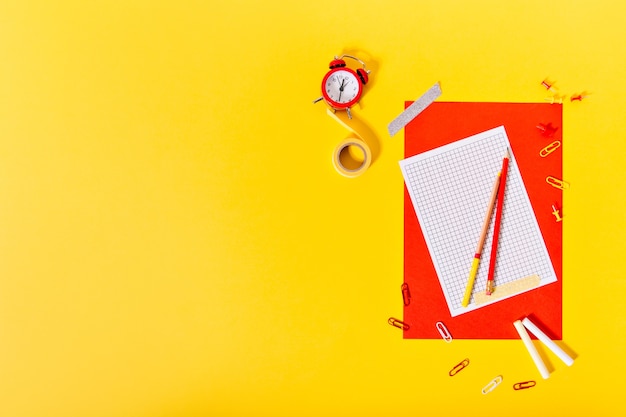 Photo d'une feuille de papier rouge, crayons, ruban adhésif et réveil sur mur jaune