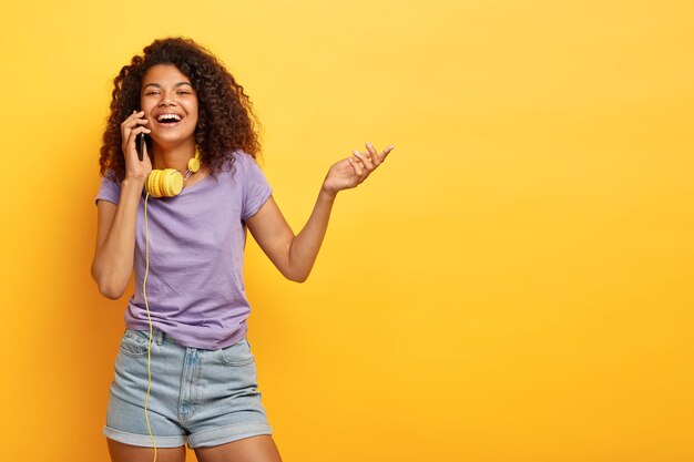 Photo d'une fille afro-américaine positive parle sur téléphone mobile, sourit largement, lève la main, partage des impressions sur le shopping, discute des dernières tendances de la mode