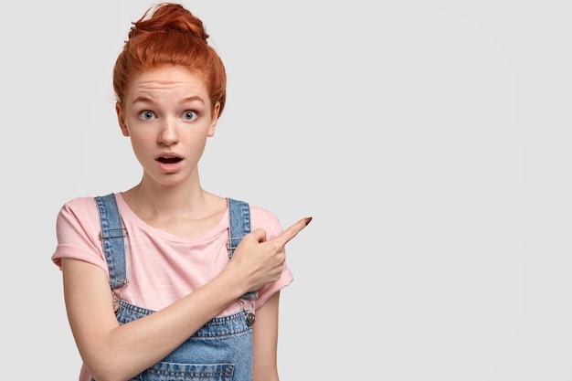 La Photo D'une Fille Aux Taches De Rousseur Aux Cheveux Rouges étonnée Indique Avec L'index Dans Le Coin Supérieur Droit, Garde La Mâchoire Tombée De Stupeur,