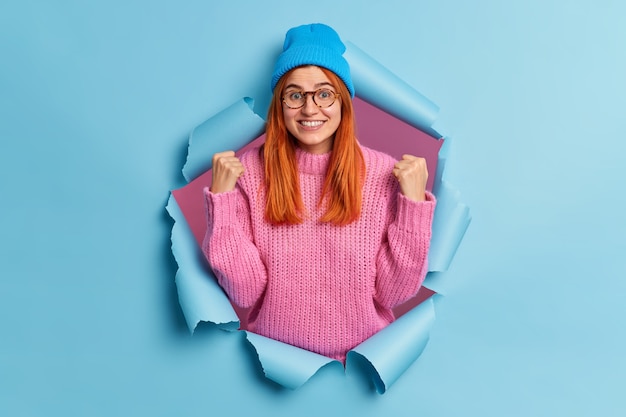 Photo d'une fille du millénaire rousse ravie lève les poings fermés célèbre le succès sourit porte largement un chapeau bleu et un pull en tricot rose perce le trou de papier