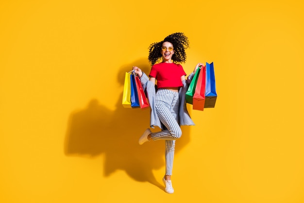 Photo d'une fille folle tenir les paquets du magasin porter un t-shirt rouge jeans rayé veste lunettes de soleil baskets isolées fond de couleur jaune