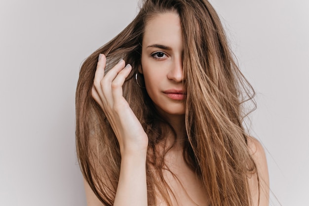 Photo en gros plan de l'adorable jeune femme touchant doucement ses cheveux. Portrait intérieur de belle femme heureuse isolée.