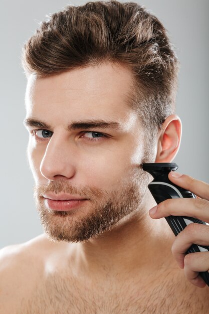 Photo gros plan d'un beau mec adulte se toilettant le visage en se rasant la joue à l'aide d'un rasoir électrique sur un mur gris