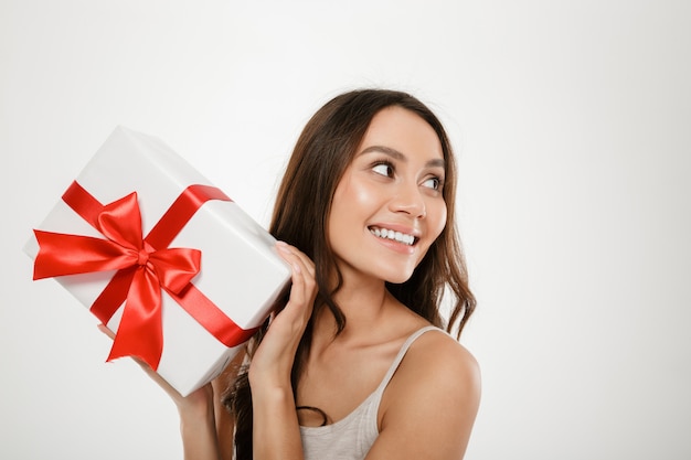 La photo en gros plan de femme caucasienne ravie à la recherche de côté tout en montrant la boîte-cadeau avec ruban rouge sur l'appareil photo, isolé sur blanc