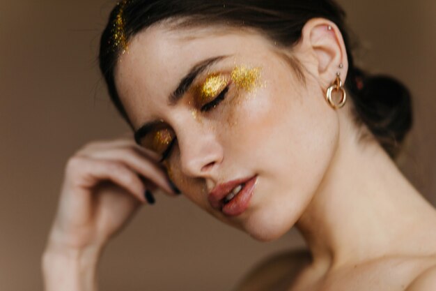 Photo en gros plan d'une fille endormie avec du maquillage de fête. Portrait intérieur de jolie femme aux cheveux noirs posant sur un mur sombre.