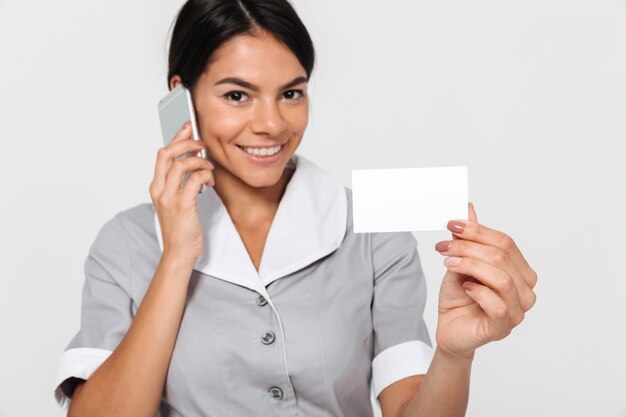 Photo en gros plan de l'heureuse jeune femme de chambre en uniforme gris parlant au téléphone tout en montrant la carte de signe vide