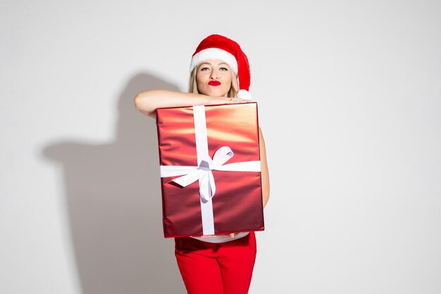 Photo en gros plan d'une jeune femme blonde portant un bonnet de Noel et un rouge à lèvres tout en s'appuyant sur un cadeau de Noël. Concept de vacances