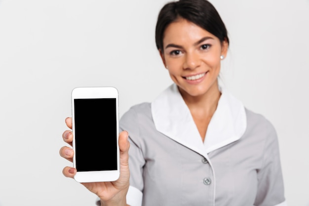 Photo en gros plan de jeune femme séduisante en uniforme montrant un écran mobile vierge, mise au point sélective sur l'affichage