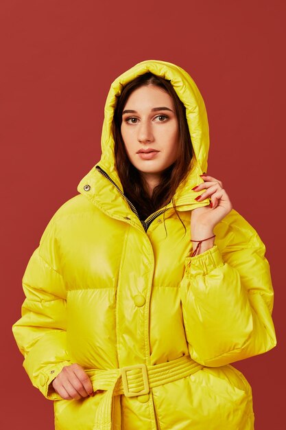 Photo en gros plan d'une jolie fille aux cheveux noirs en studio, en veste jaune vif. Fond rouge, vêtements oversize, capuche