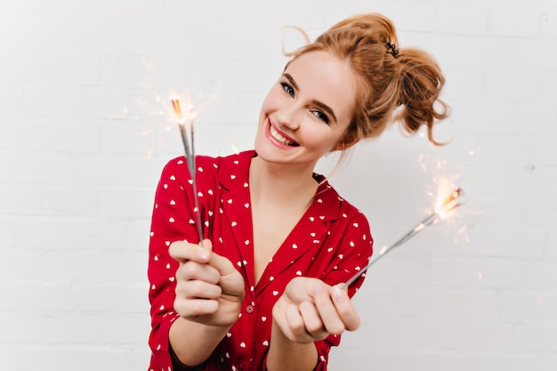 Photo en gros plan de joyeuse fille caucasienne tenant des lumières du Bengale. Portrait de jeune femme heureuse en vêtements de nuit rouge isolé sur un mur blanc avec des cierges magiques.