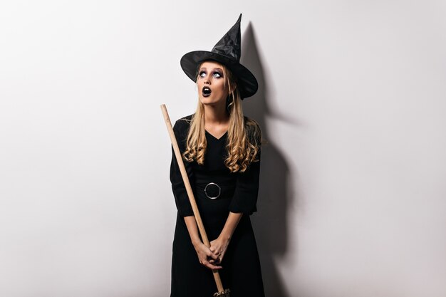 Photo d'Halloween de jolie fille blonde avec un balai magique. Plan intérieur d'une jeune sorcière curieuse au chapeau noir.