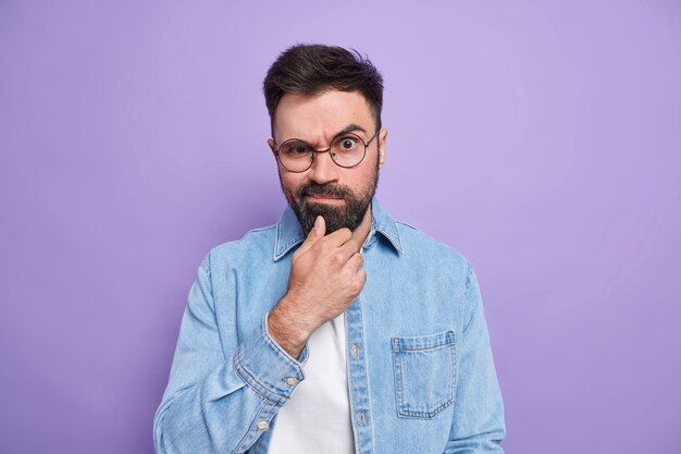 Photo d'un homme adulte barbu sérieux tient le menton lève les sourcils fronce les sourcils le visage vêtu d'une chemise à la mode étant insatisfait de quelque chose