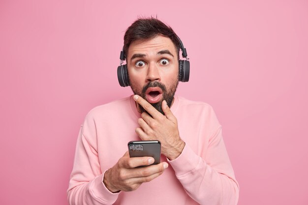 La photo d'un homme adulte barbu surpris regarde avec une expression étonnée tient le menton écoute un livre audio ou de la musique préférée via un casque sans fil habillé avec désinvolture réagit émotionnellement à une offre inattendue