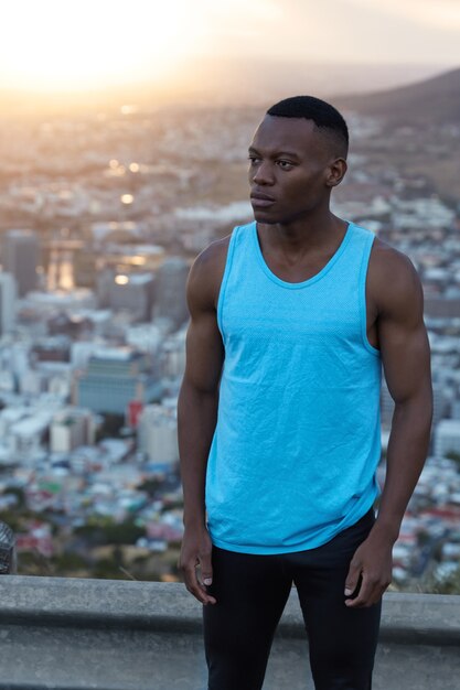 Photo d'un homme athlétique motivé à la peau foncée avec des muscles, s'entraîne régulièrement en plein air, pense à un nouveau défi, porte un t-shirt bleu et des leggings noirs, se dresse contre la vue sur la ville avec le soleil.