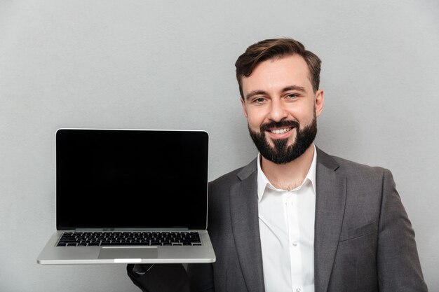 Photo d'homme barbu satisfait tenant un carnet d'argent démontrant ou faisant de la publicité sur l'appareil photo, isolé sur un mur gris