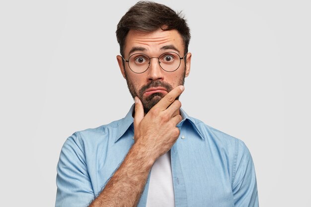 Photo d'un homme barbu surpris tient le menton et regarde avec hésitation, s'interroge sur les dernières nouvelles, porte des lunettes et une élégante chemise bleue, isolée sur un mur blanc. Concept de personnes et d'expressions faciales