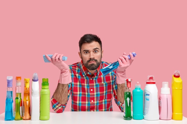 Photo d'un homme barbu vêtu d'une chemise à carreaux, utilise différents produits de nettoyage, entouré de bouteilles en plastique, a l'air étonnamment