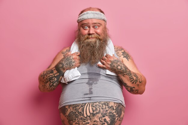 Photo d'un homme drôle aux yeux bleus a un gros ventre, un corps tatoué, occupé à faire des exercices de fitness, brûle des calories après avoir mangé de la restauration rapide, isolé sur un mur rose. Un homme de grande taille mène un mode de vie sain