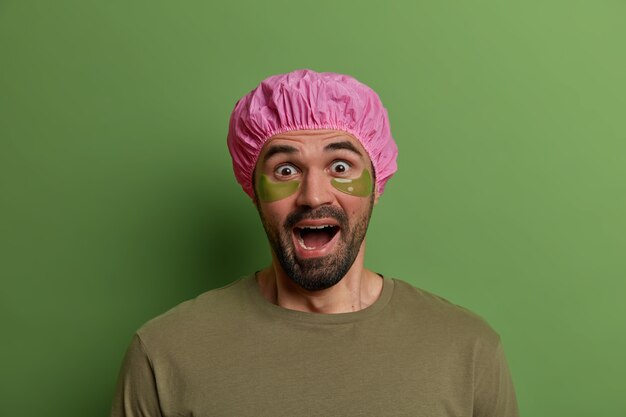 Photo d'un homme effrayé et émotionnel regarde avec les yeux sortis et hurle de peur, applique des tampons de collagène sous les yeux, reçoit des soins de beauté, porte un chapeau de bain protecteur, se tient à l'intérieur. Bien-être, beauté