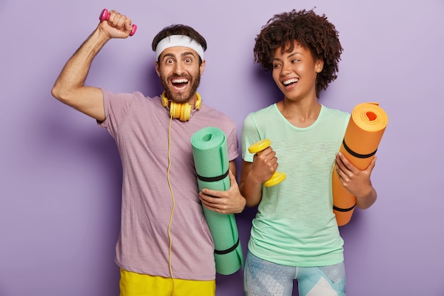 Photo d'un homme et d'une femme heureux travaillent sur des biceps avec des poids, portent des karemats, ont des expressions joyeuses, s'entraînent ensemble, portent des vêtements décontractés, sont motivés pour un mode de vie sain et le sport