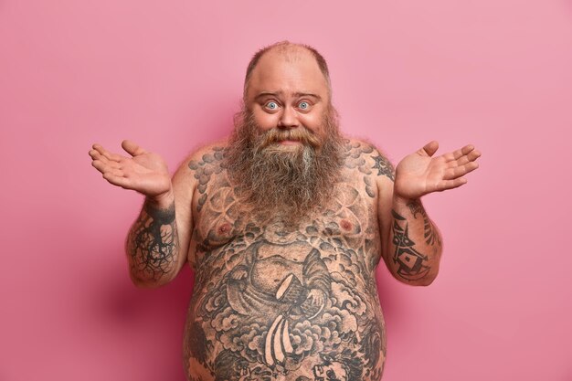 La photo d'un homme gai hésitant écarte les paumes sur le côté, dit que je ne sais pas, être heureux et confus, a un gros ventre, un corps tatoué, ne sait pas comment être en forme et perdre du poids, isolé sur un mur rose