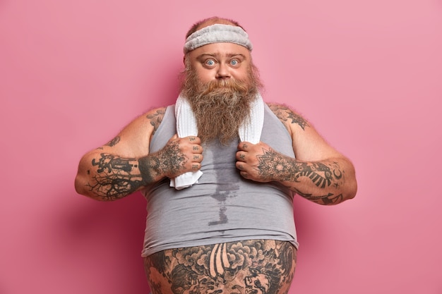 Photo gratuite photo d'un homme gros et sérieux en tenue de sport rêve de corps musclé, travaille dur sur son corps, veut perdre du poids, a les bras tatoués, gros ventre, fait des exercices physiques avec un entraîneur de fitness