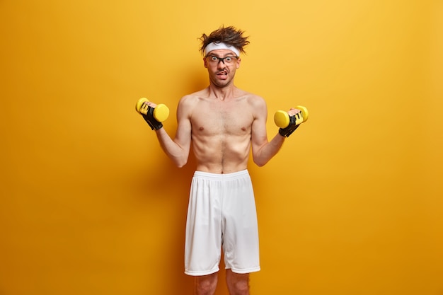Photo d'un homme maigre fait du sport, développe ses muscles à la maison, a un complexe d'entraînement efficace avec des haltères, porte un short blanc, pose avec le torse nu contre un mur jaune. Concept de soins de santé